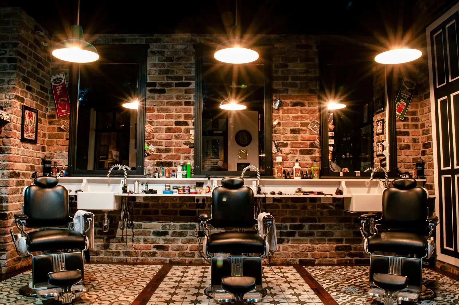 Three leather barber chairs facing mirrors on a brick wall under round lamps