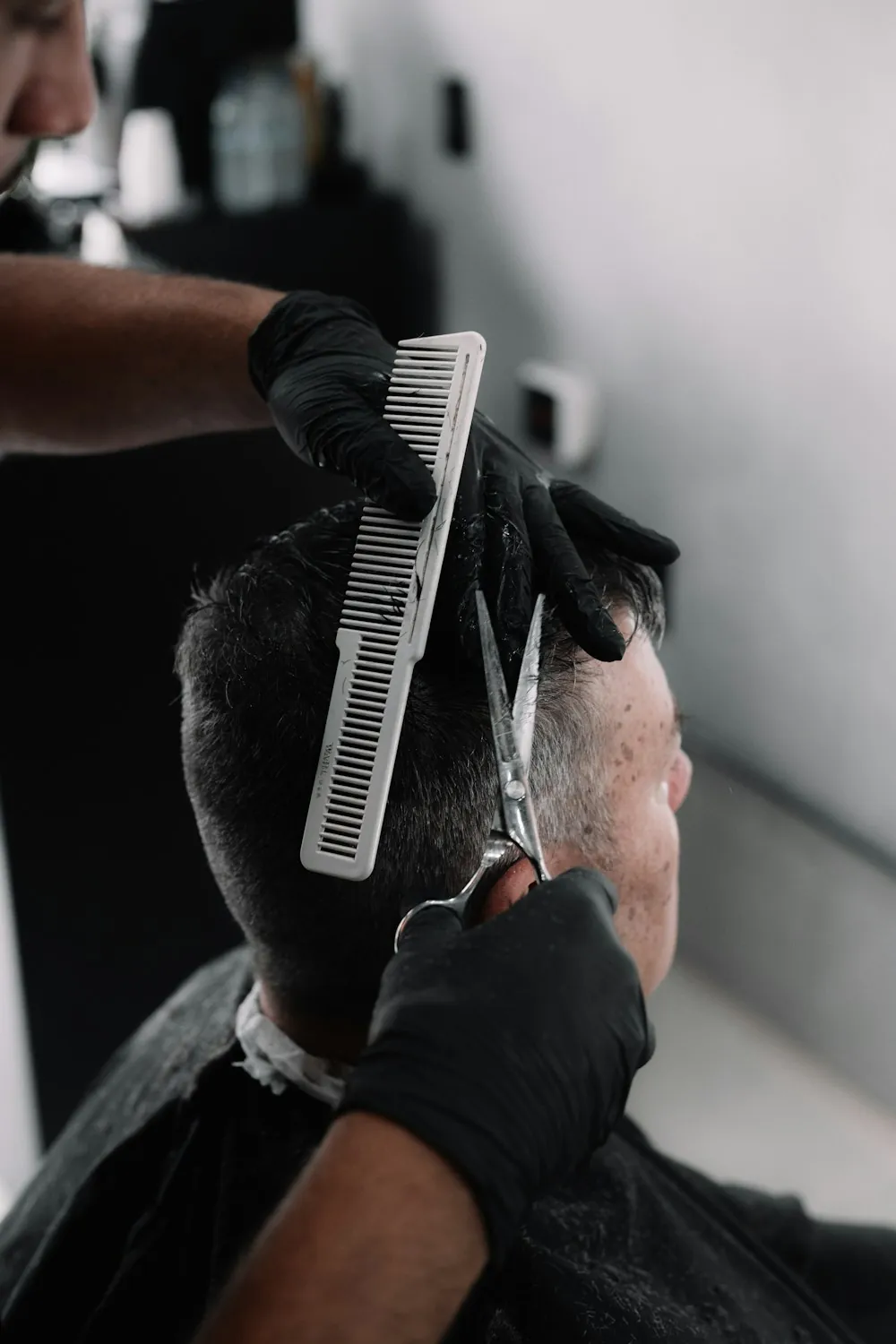A barber in black gloves combing and cutting a client's hair, seen from behind