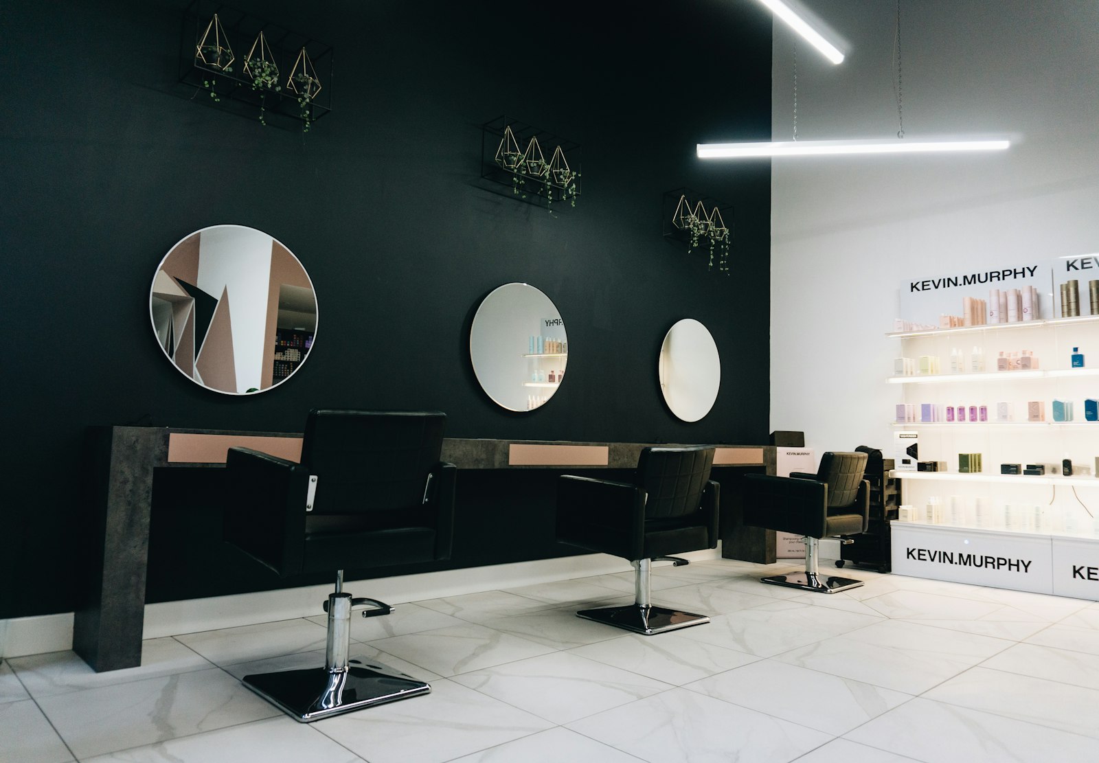 Salon stations with round mirrors on a dark wall and black chairs on a white tile floor